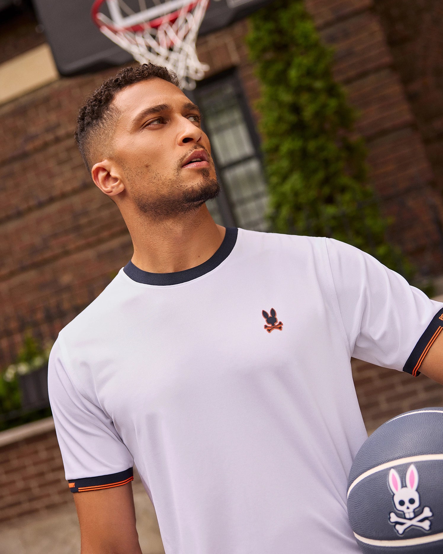 A man wearing the Psycho Bunny MENS EDMONDS SPORT TEE - B6U857D200, a quick-drying white T-shirt with a small bunny logo, stands outdoors holding a basketball. He is looking to his right, with a basketball hoop visible in the background. The setting appears to be urban with a brick wall and greenery.