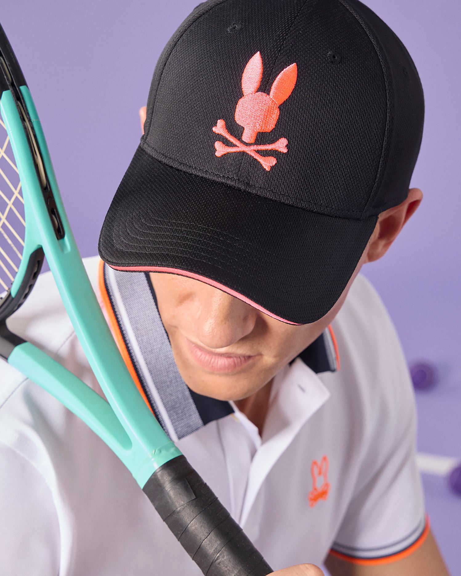 A person wearing a Psycho Bunny MENS DOVER SPORT CAP with a pink bunny logo, partially visible, holding a tennis racket close to their face. They are dressed in a white polo with contrasting orange and gray trim.