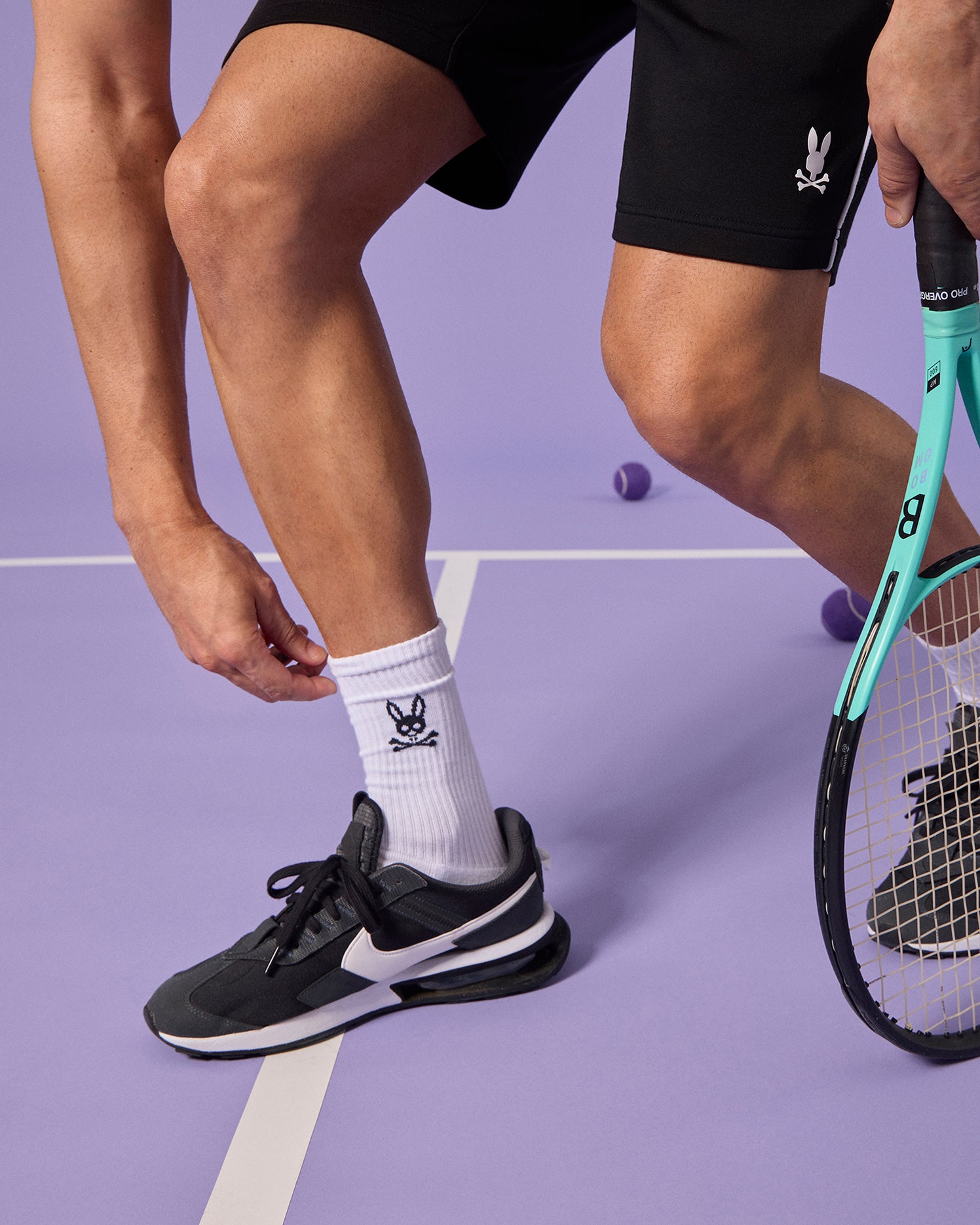 A close-up shot of a person's lower legs and feet, wearing black sneakers and Psycho Bunny men's sport socks with an Embroidered Bunny Logo, standing beside a green tennis racquet on a purple court.