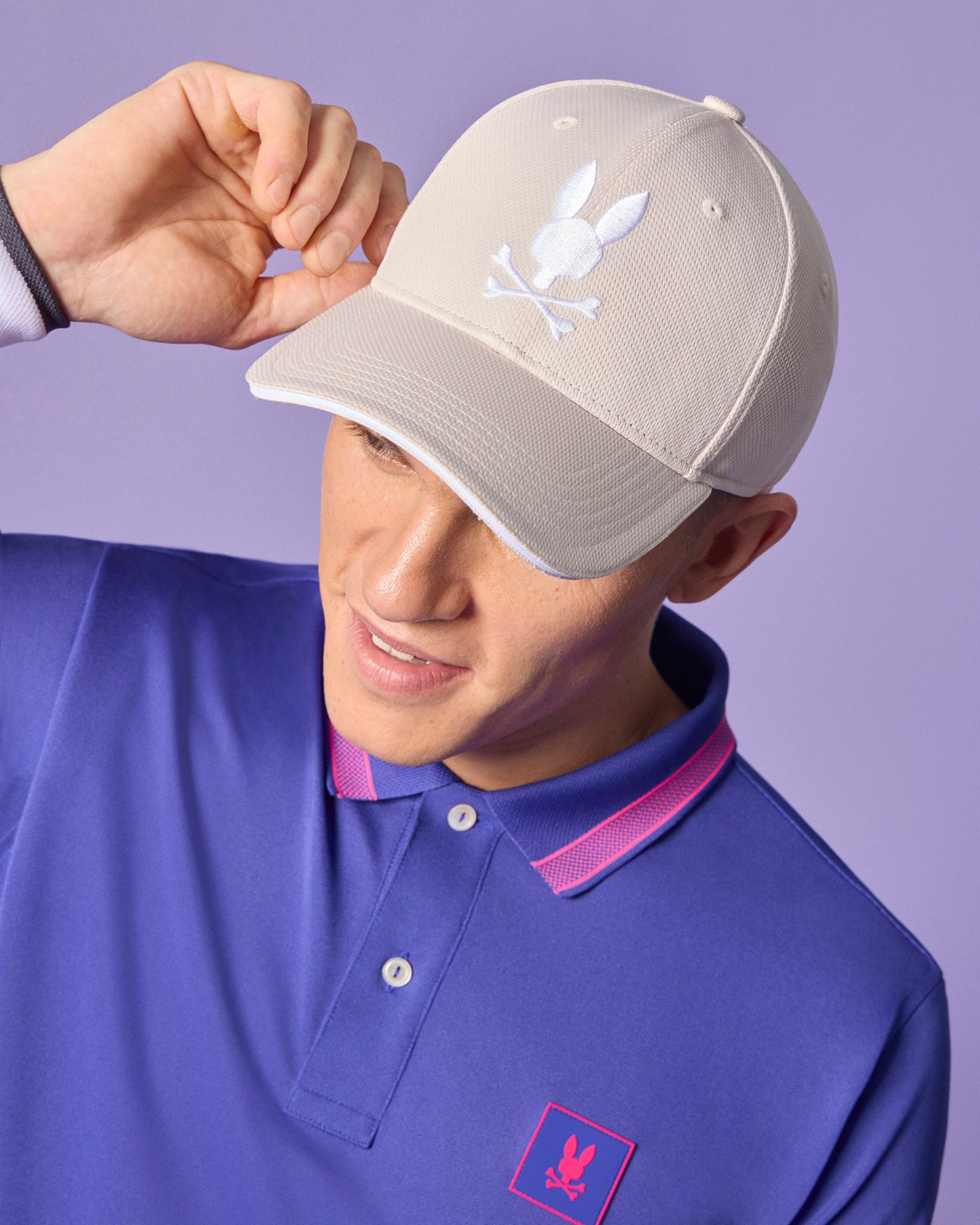 A young man in a blue polo shirt playfully tips his Psycho Bunny Men's Dover Sport Cap - B6A285B2HT, adorned with embroidered white rabbit logos, against a complementary purple background.
