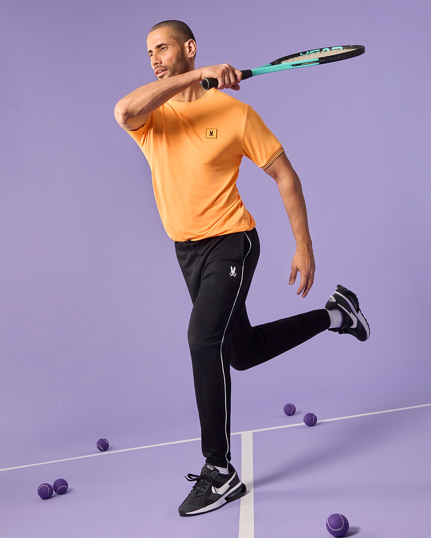 A person wearing a Psycho Bunny MENS TARRYTOWN SPORT TEE - B6U287B200 in breathable stretch jersey and black pants plays tennis on a lavender court. They are in a mid-swing position with their left foot off the ground. Several tennis balls are scattered on the court, and the person's moisture-wicking shirt keeps them cool as they hold a racket in their right hand.
