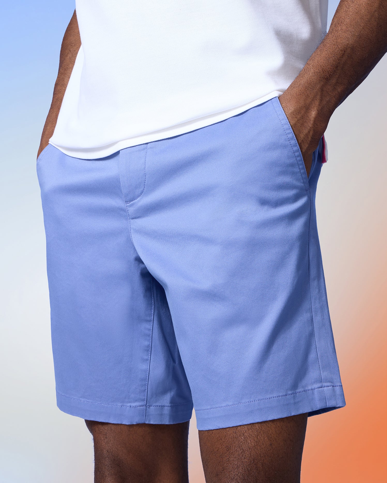 A person stands with their hands in the pockets of vibrant blue Psycho Bunny MENS YORK CHINO SHORT - B6R357B200, wearing a white t-shirt. The background features a gradient of pastel colors, blending from blue to orange.