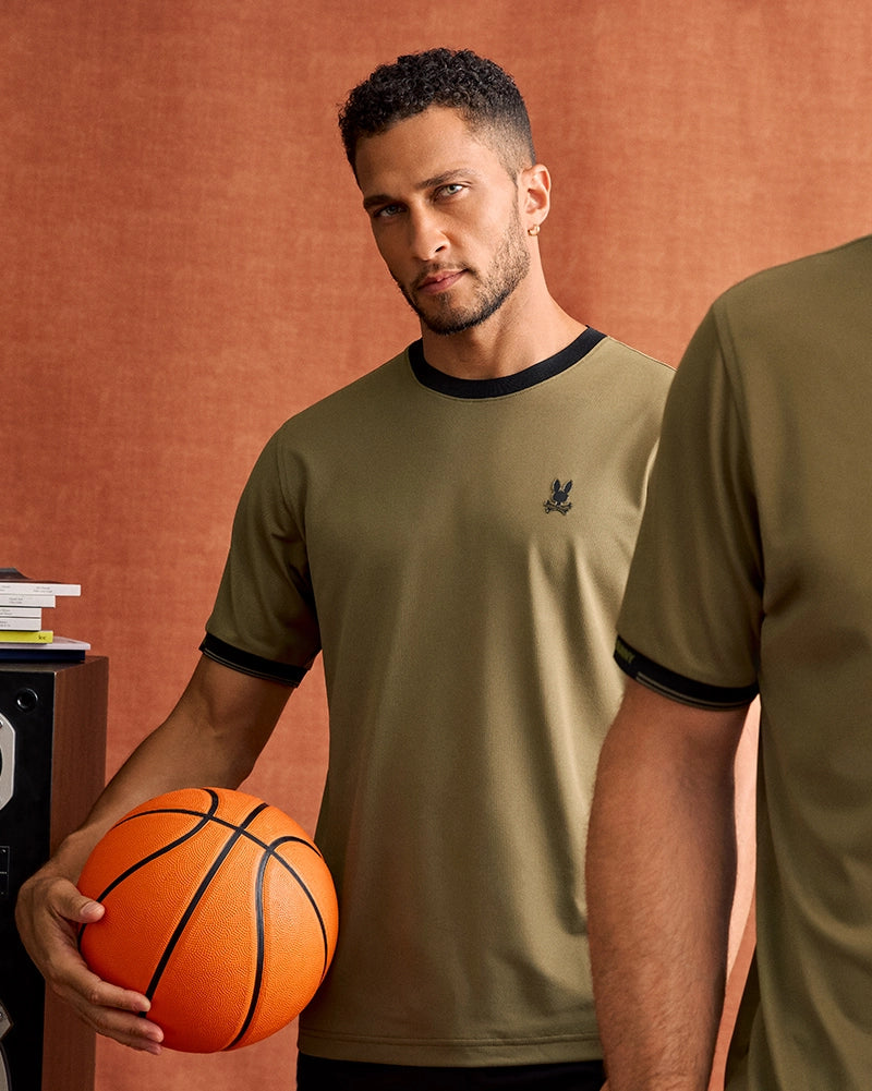 A man with short curly hair and a trimmed beard is holding a basketball in one hand. He is wearing the Psycho Bunny MENS EDMONDS SPORT TEE - B6U857D200, a khaki-colored, quick-drying T-shirt with black trim and an embroidered emblem on the chest. The background features a warm, orange-brown backdrop and part of another person in similar attire.