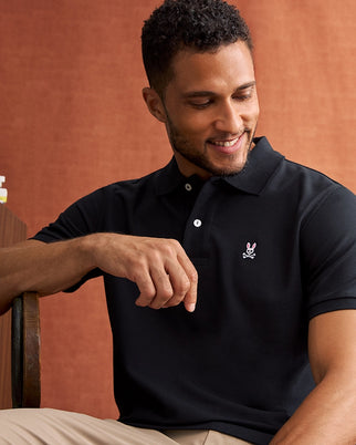 A smiling man with short hair sits in a chair, wearing a Psycho Bunny Men's Classic Pique Polo Shirt (B6K001ARPC) made of 100% Pima cotton. The black shirt features a small embroidered logo of the Psycho Bunny bunny above crossed bones on the left side of the chest and is adorned with mother-of-pearl buttons. He rests one arm on the back of the chair against a warm, textured brown background.