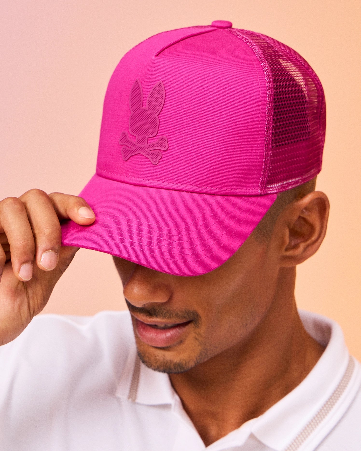 A person wearing a white polo shirt tilts the brim of a bright pink baseball cap adorned with a bunny head and crossbones logo. The Mens Riviera Trucker Cap - B6A667C200 by Psycho Bunny, with its iconic snapback fastening, evokes early 2000s nostalgia against a soft gradient of pinks and peaches.