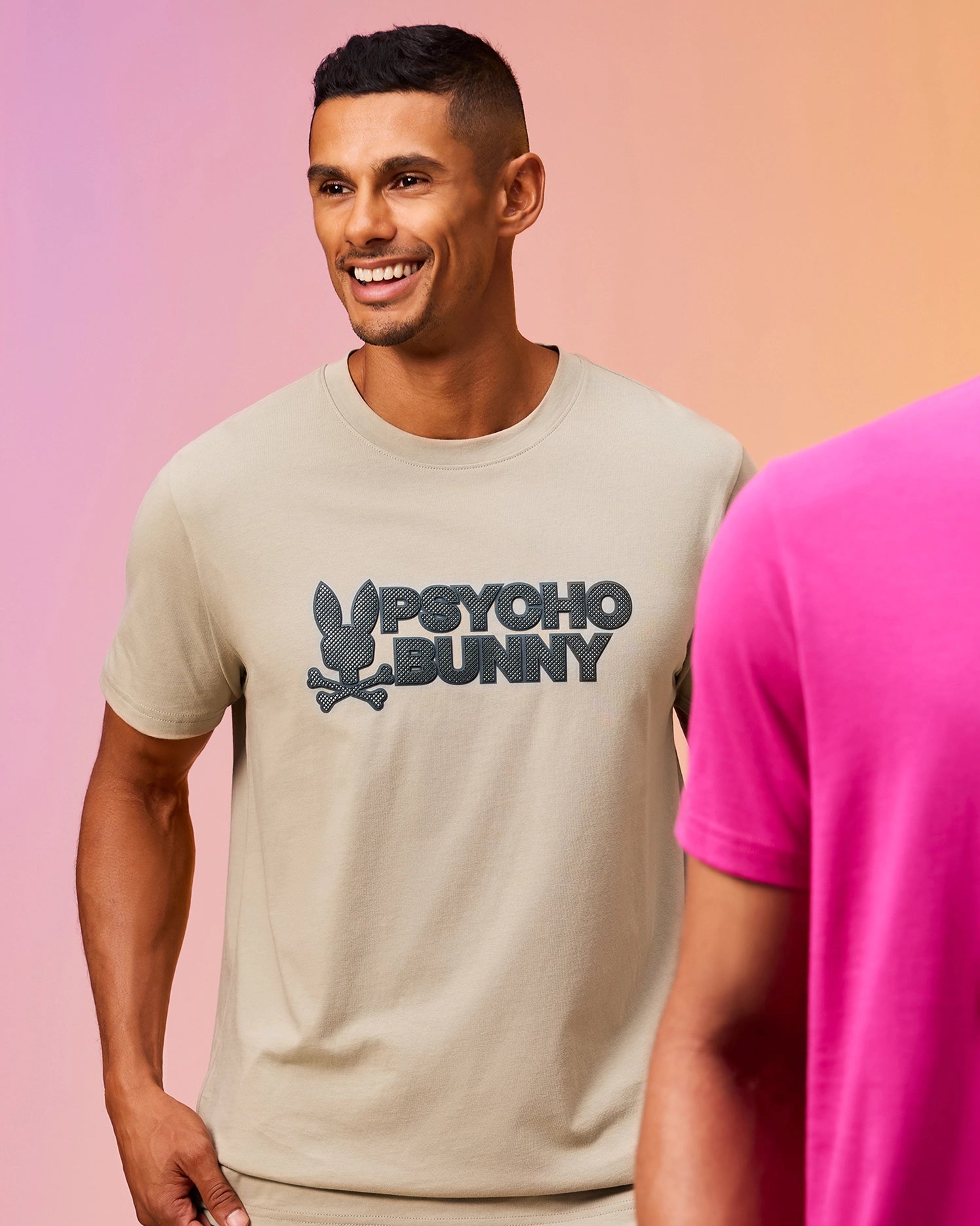 A man with short hair is smiling and wearing a light beige MENS MELVIN GRAPHIC TEE - B6U663C200 by Psycho Bunny, made of Pima cotton, featuring a large logo of a bunny and the text 
