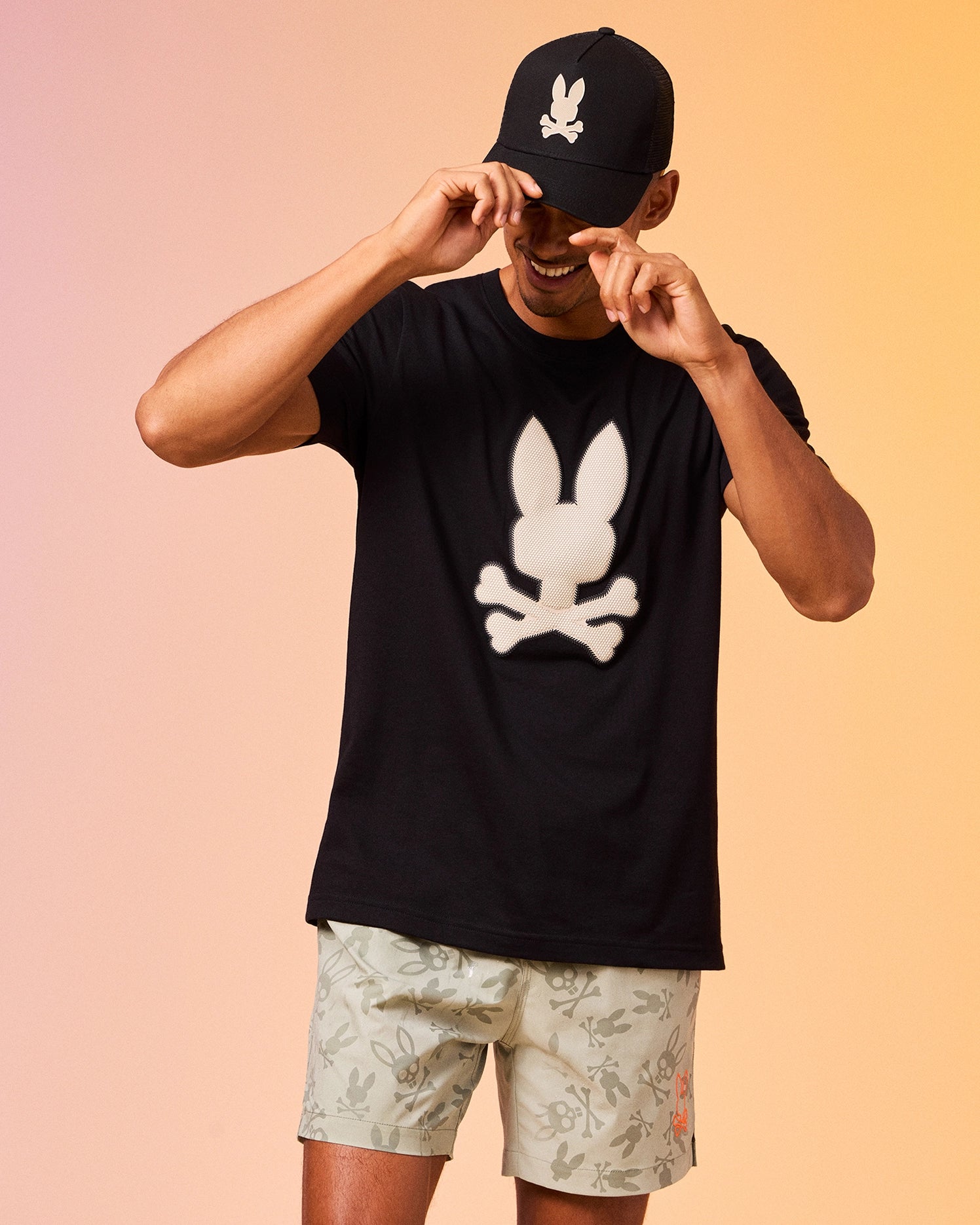 A person wearing a black cap and a Psycho Bunny MENS RIVIERA GRAPHIC TEE - B6U639C200 with a skull and rabbit ears design. They are also wearing light-colored shorts with a pattern. The person is adjusting their cap while smiling. The background has a gradient of light pink and orange colors.