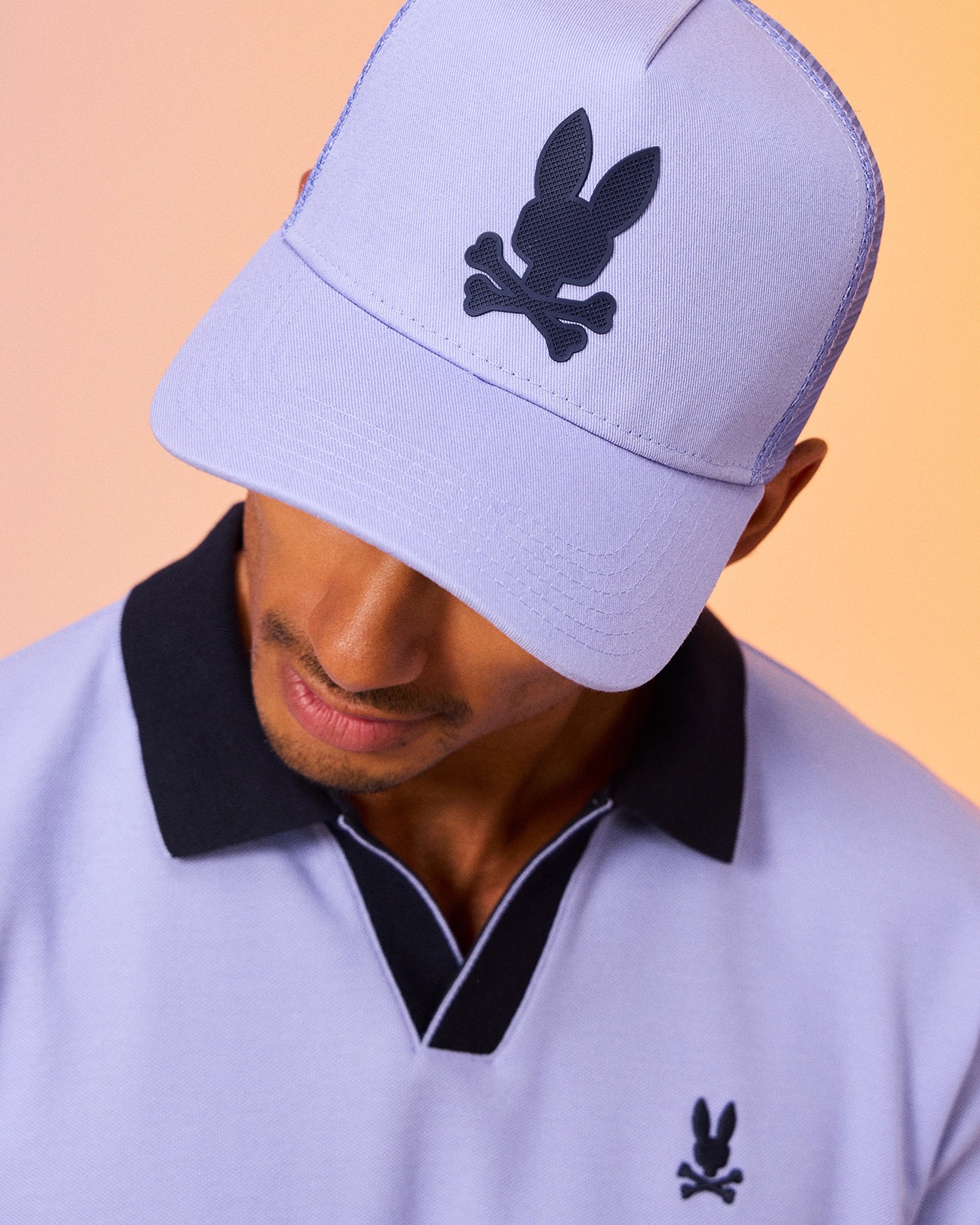 A man wearing a light purple Psycho Bunny MENS RIVIERA TRUCKER CAP - B6A667C200 with a black bunny and crossbones logo looks down. He is also dressed in a matching light purple polo shirt featuring the same logo. The background has a warm gradient, creating a stylish and cohesive look with early 2000s nostalgia.