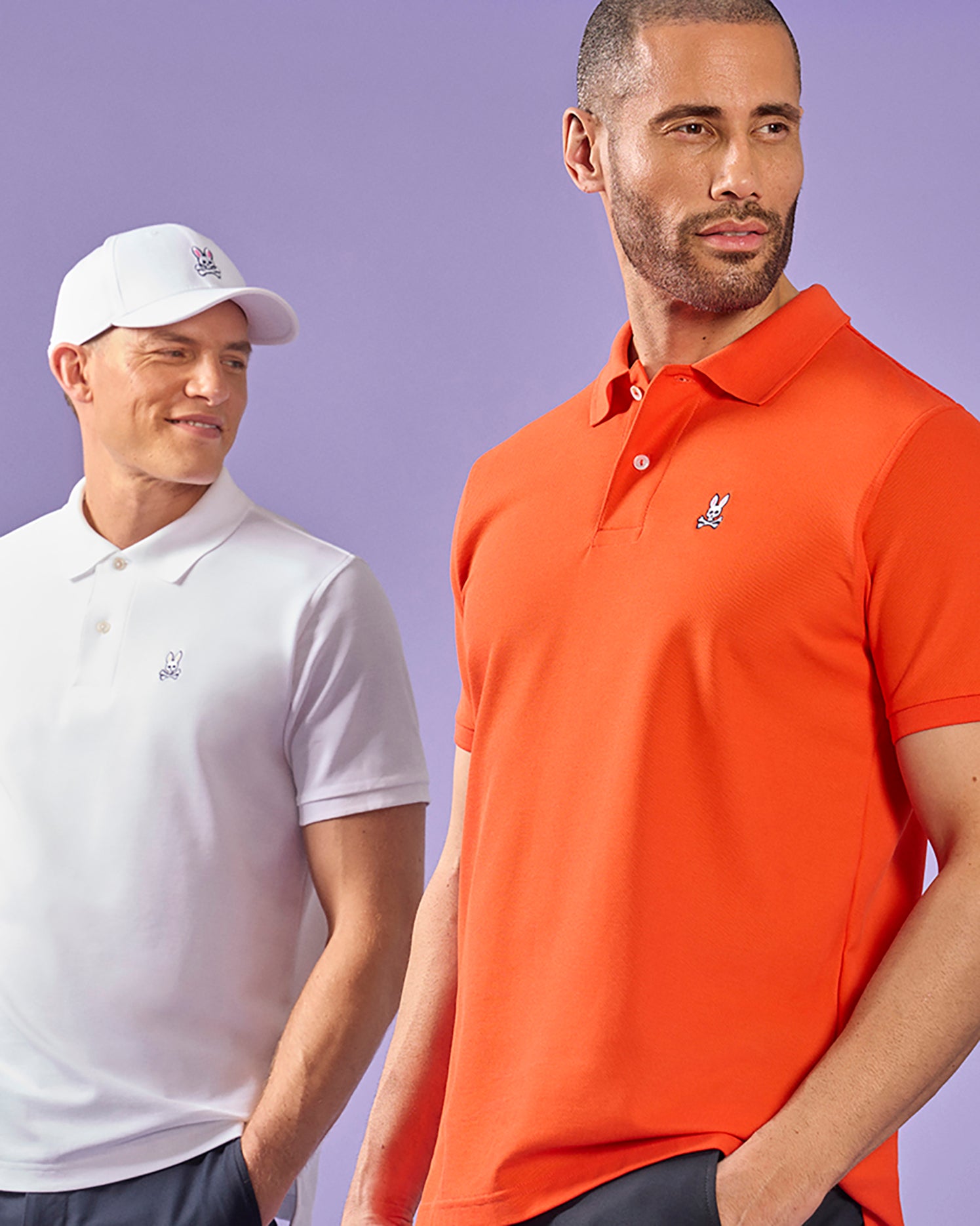 Two men modeling Psycho Bunny classic piqué polo shirts, one in white and the other in orange, against a dual-tone purple background. Both shirts feature a small Psycho Bunny logo on the chest.
