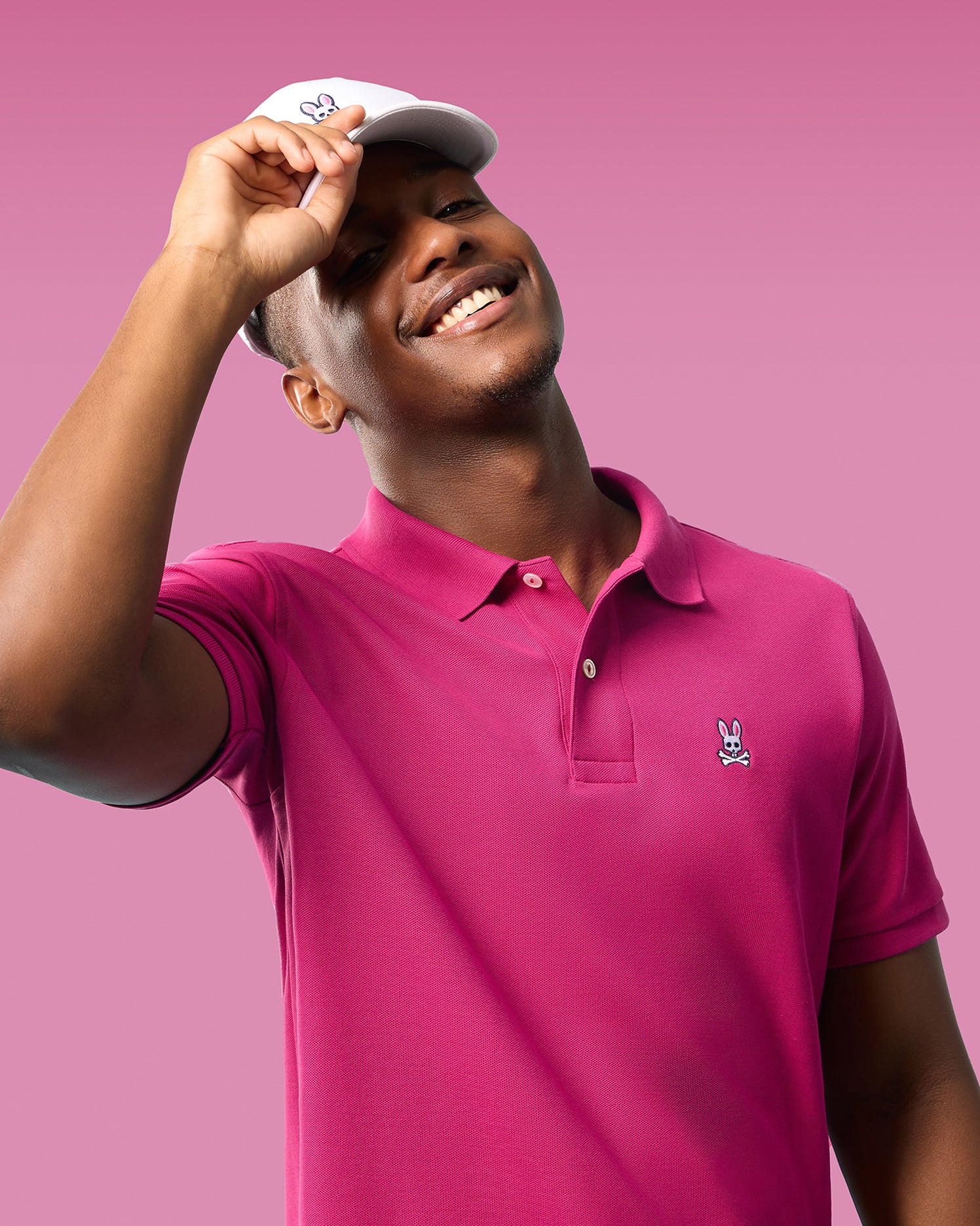 A smiling young man tilting a white cap with a Psycho Bunny logo, wearing a pink Psycho Bunny Pima cotton polo shirt (MENS CLASSIC POLO - B6K001CRPC) with a matching bunny emblem, against a pink background.