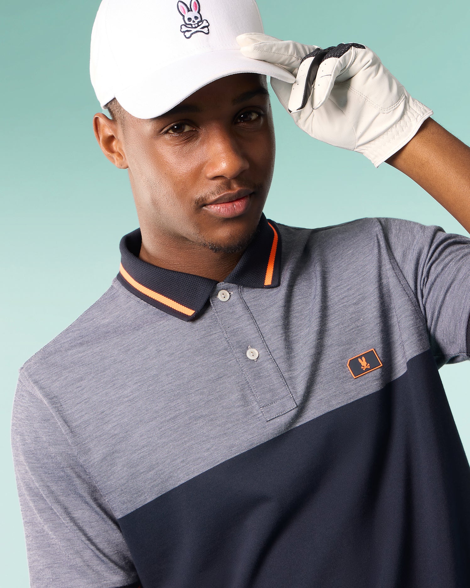 A man wearing a Psycho Bunny MENS SYRACUSE SPORT POLO SHIRT - B6K341B200 with a gray top and black bottom, complemented by an orange trim on the collar, holds the brim of his white cap adorned with a pink rabbit logo. He is also wearing a white golf glove and stands against a light teal background.