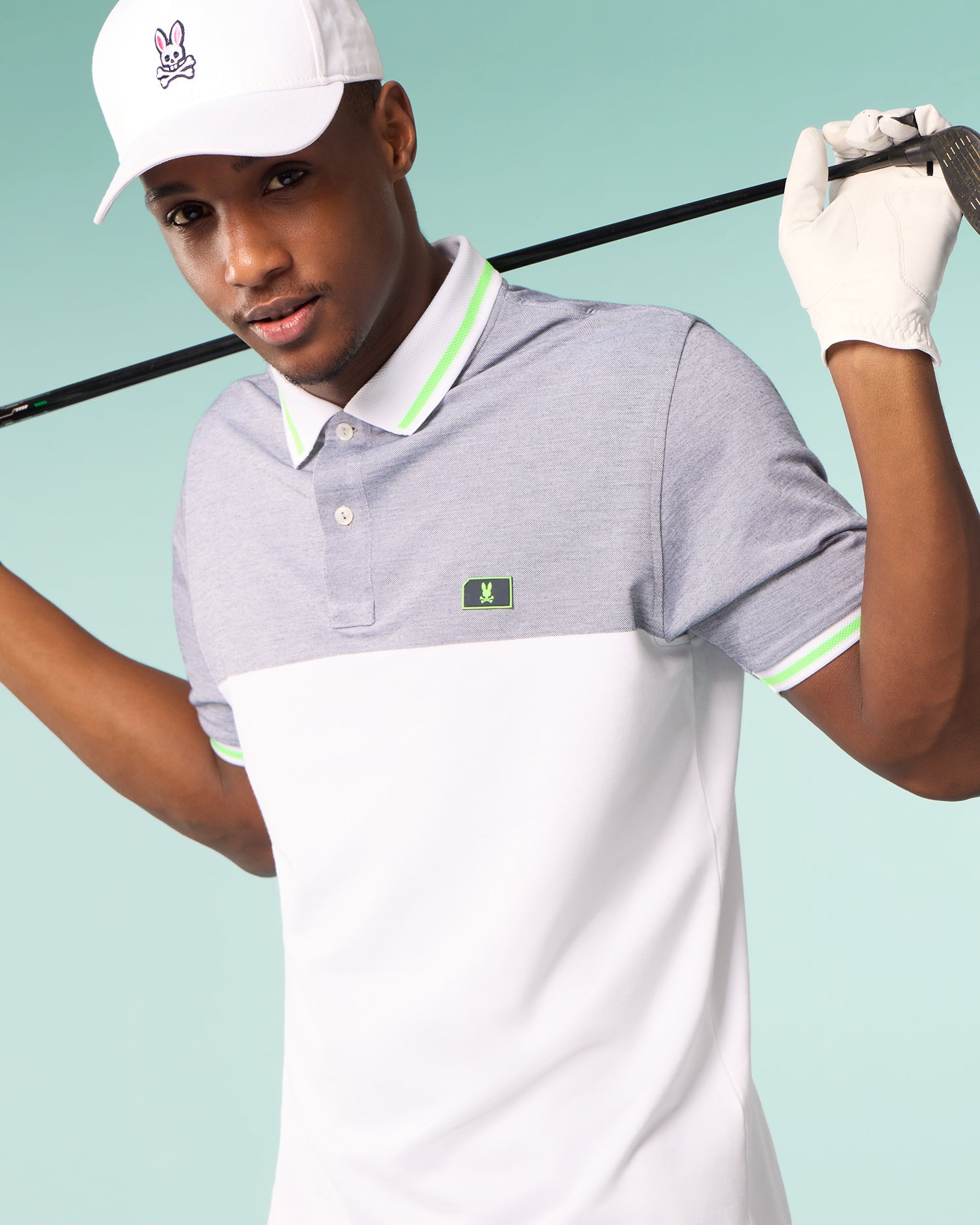 A man in a Psycho Bunny MENS SYRACUSE SPORT POLO SHIRT - B6K341B200 with green accents and a small green logo stands holding a golf club behind his neck. He also wears a white cap with a pink bunny logo and a white golf glove on one hand. The background is a solid light teal color.