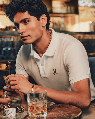 Dressed in a MENS ALEXANDER PIQUE POLO by Psycho Bunny, known for its durable breathability, a person sits at a poker table holding cards. Poker chips are scattered across the table, and a glass of liquor is prominently placed in the foreground. The background is warmly lit, creating a cozy atmosphere.