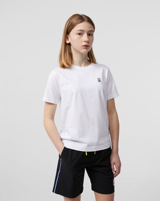 A person with shoulder-length light brown hair wearing a plain white Psycho Bunny Kids Classic Crew Neck Tee - B0U014CRPC and black shorts stands against a plain background with hands in pockets. The 100% Pima cotton t-shirt features a small embroidered bunny logo on the left chest, perfect for back to school outfits.