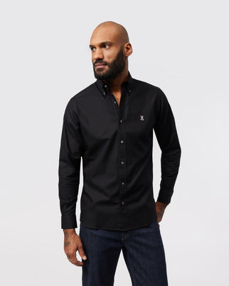 A man with a beard and shaved head is standing against a plain backdrop, wearing a black long-sleeve MENS CLASSIC OXFORD LONG SLEEVE SHIRT - B6C655X1PC by Psycho Bunny with an embroidered Bunny logo on the chest and dark blue jeans. He has his left hand in his pocket and is looking to the side.