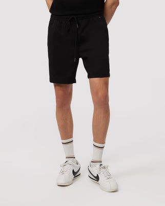 A man in Psycho Bunny eco-friendly shorts and white sneakers with black accents, standing on a plain background. The focus is on the lower half of his body from the waist down.