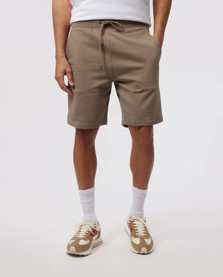 A person wearing beige Psycho Bunny FRENCH TERRY SHORTS, white socks, and beige sneakers. Only the lower half of the figure is visible against a plain white background.