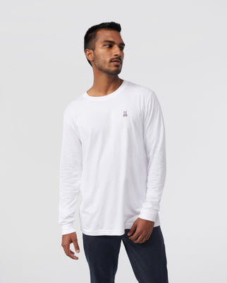 A person is wearing the Psycho Bunny MENS LONG SLEEVE CLASSIC CREW NECK TEE - B6T422ARPC, which is white with a small embroidered bunny logo on the chest. They pair it with dark pants and stand against a plain light gray background.