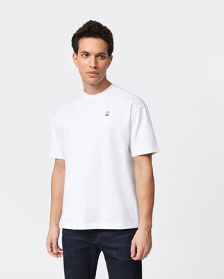 A man with short dark hair is wearing a plain white MENS OVERSIZED CLASSIC CREW NECK TEE - B6U240W1PC from Psycho Bunny and dark jeans, standing against a plain white background. The heavyweight fit t-shirt has a small embroidered logo on the left side of the chest. The man is looking slightly to the side with a neutral expression.