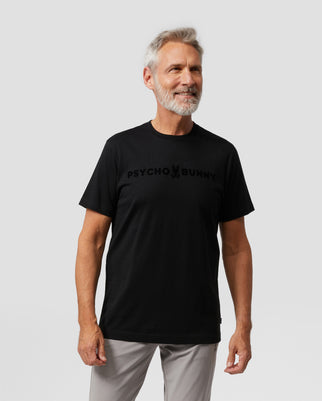 A smiling older adult with gray hair and a beard wears the Psycho Bunny MENS ROYCE FLOCKING GRAPHIC TEE (B6U248W1PC) made from soft Pima cotton, paired with light gray pants. He stands against a plain light gray background, highlighting the shirt's high-pile brushed logo.