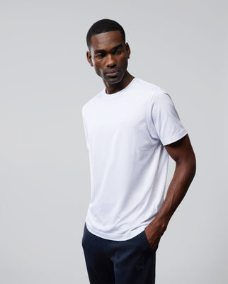 A man stands against a plain background, wearing a white MENS OUTLINE TEE - B6U500ARPC from Psycho Bunny and dark pants. His right hand is in his pocket while his left arm hangs by his side. He looks slightly to his right with a relaxed expression.